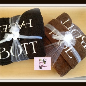 May include: Two brown towels wrapped in white tulle with white embroidery that reads "Face" and "Butt".