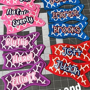 May include: A collection of colorful fabric can coolers with names embroidered on them. The can coolers are shaped like mermaid tails and have a scale pattern. The text "Offset Mermaid Tails font" and "Offset Shakeitoff Font" is at the top of the image. The text "Tini's Sewing and Embroidery" is at the bottom of the image.