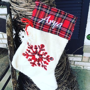 May include: White Christmas stocking with a red and black plaid cuff. The stocking has a red snowflake design and the name "Arya" embroidered on the cuff.