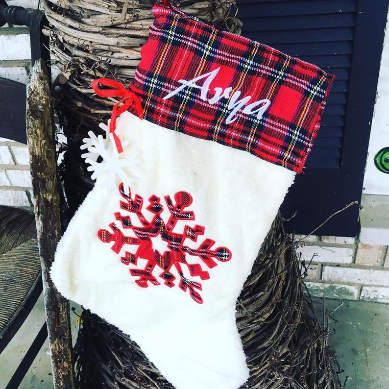 May include: White Christmas stocking with a red and black plaid cuff. The stocking has a red snowflake design and the name "Arya" embroidered on the cuff.