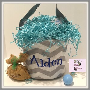 May include: A gray and white chevron fabric Easter basket with a blue handle. The basket is filled with blue shredded paper and has the name "Aiden" embroidered on the front. A small brown bunny and a blue Easter egg are in front of the basket.