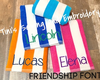 May include: Four striped beach towels with personalized names embroidered in different colors. The names are "Tini's", "Lincoln", "Lucas", and "Elena". The towels are white with blue, orange, pink, and blue stripes. The text "Friendship Font" is at the bottom of the image.