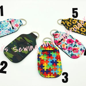 K&ouml;nnte beinhalten: F&uuml;nf verschiedene Handdesinfektionsmittel-Halter mit Schl&uuml;sselanh&auml;ngern. Die Halter sind aus Stoff und haben verschiedene Muster, darunter Blumen, Tarnmuster, Puzzle-Teile, Sonnenblumen und ein Blumenmuster mit rosa Bl&uuml;ten. Die Halter sind von 1 bis 5 nummeriert.