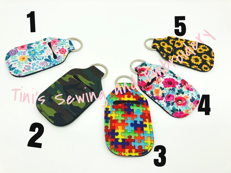 K&ouml;nnte beinhalten: F&uuml;nf verschiedene Handdesinfektionsmittel-Halter mit Schl&uuml;sselanh&auml;ngern. Die Halter sind aus Stoff und haben verschiedene Muster, darunter Blumen, Tarnmuster, Puzzle-Teile, Sonnenblumen und ein Blumenmuster mit rosa Bl&uuml;ten. Die Halter sind von 1 bis 5 nummeriert.