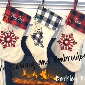 May include: Three white Christmas stockings with red and black buffalo plaid trim, personalized with the names "Big Pop", "Rose", and a snowflake design. The stockings are hanging on a white mantel above a fireplace. The text "Tini's Sewing and Embroidery" and "Berkley Font" is written below the stockings.