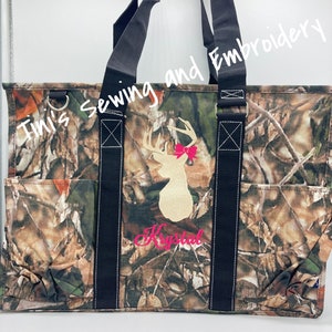 May include: A camouflage tote bag with black straps and a white deer with a pink bow embroidered on the front. The bag has a pocket on the side and the text "Timi's Sewing and Embroidery" and "Krystal" are embroidered on the bag.