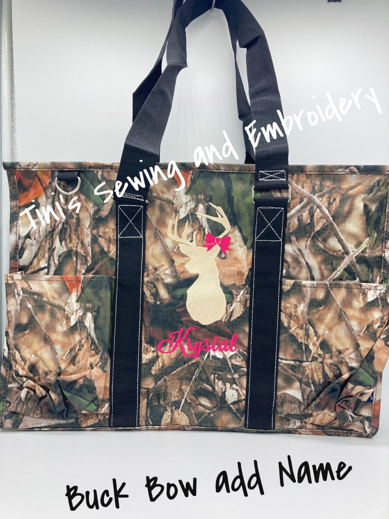 May include: A camouflage tote bag with black straps and a white deer with a pink bow embroidered on the front. The bag has a pocket on the side and the text "Timi's Sewing and Embroidery" and "Krystal" are embroidered on the bag.
