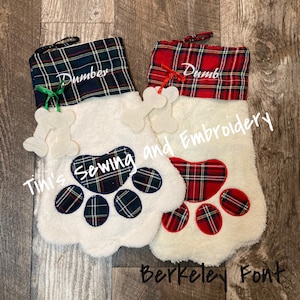 May include: Two white plush Christmas stockings with red and green plaid fabric at the top. The stockings have a paw print design and are personalized with the names "Dumber" and "Dumb". The stockings are decorated with white bone ornaments. The text "Tini's Sewing and Embroidery" and "Berkeley Font" is visible in the image.