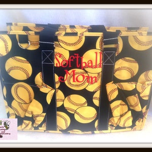 Peut inclure: Un sac fourre-tout noir orné de motifs de balles de softball jaunes. Le sac a des poignées et des poches noires, avec les mots "Softball Mom" brodés en rouge. Le sac est conçu pour transporter des objets.