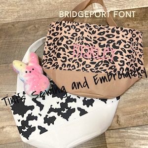 May include: Two tote bags, one with a black and white cow print and the other with a brown and black leopard print. The leopard print bag has the name "Marley" embroidered on it in pink thread. The text "Tint's Sewing and Embroidered" is visible in the image.