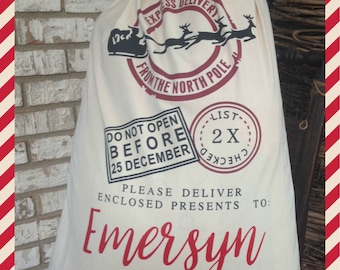 May include: A white canvas Santa sack with red and black printed text. The text reads "Express Delivery from the North Pole", "Do Not Open Before 25 December", "List 2X Checked", "Please Deliver Enclosed Presents To: Emersyn", and "If Undeliverable Return To: Santa Claus, North Pole".