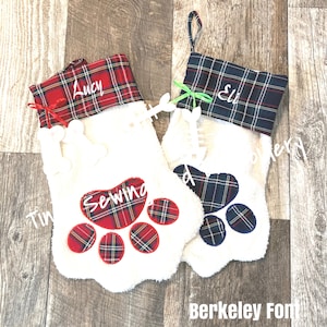 May include: Two white plush Christmas stockings with red and blue plaid fabric at the top. The stockings have a paw print design with a bone detail. The stockings are personalized with the names "Lucy" and "Eli".