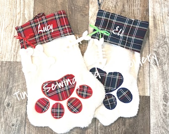 May include: Two white plush Christmas stockings with red and blue plaid fabric at the top. The stockings have a paw print design with a bone detail. The stockings are personalized with the names "Lucy" and "Eli".