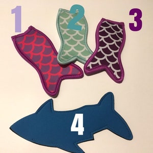 May include: Four colorful neoprene can coolers. Three are shaped like mermaid tails with purple, teal, and green scales. One is shaped like a blue shark.