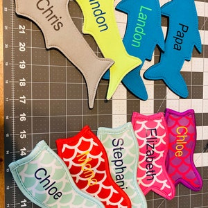 May include: A set of 9 personalized fish-shaped drink koozies. The koozies are made of neoprene and feature a variety of colors and patterns. The names "Chris", "Landon", "Papa", "Chloe", "Elizabeth", "Stephani", and "Chloe" are embroidered on the koozies.