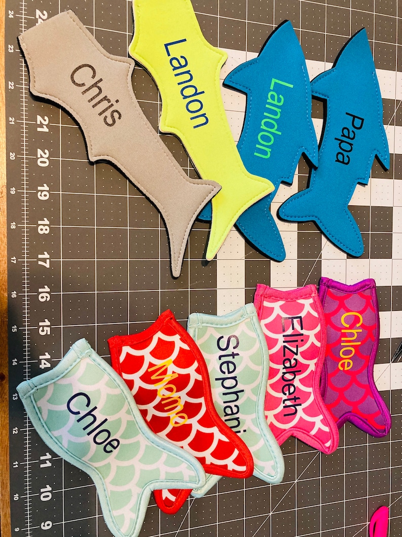 May include: A set of 9 personalized fish-shaped drink koozies. The koozies are made of neoprene and feature a variety of colors and patterns. The names "Chris", "Landon", "Papa", "Chloe", "Elizabeth", "Stephani", and "Chloe" are embroidered on the koozies.
