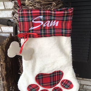 May include: Red and white plaid Christmas stocking with a white furry body and a paw print design. The stocking is personalized with the name "Sam" in white embroidery.