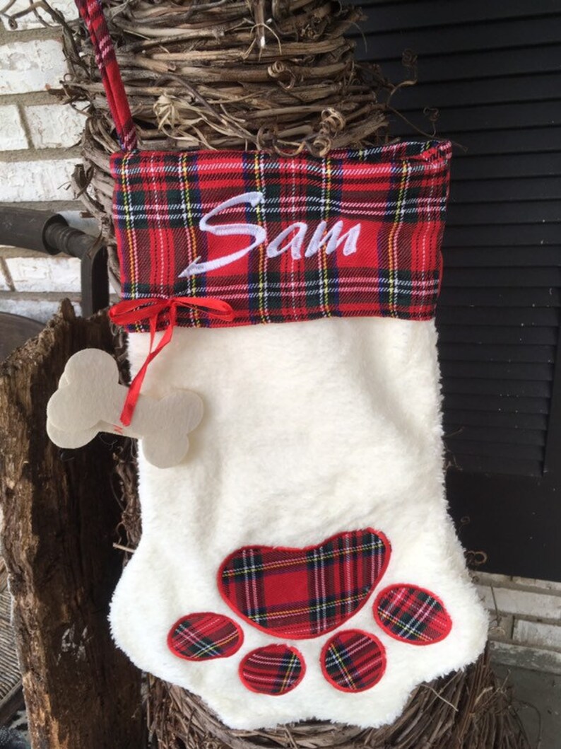 May include: Red and white plaid Christmas stocking with a white furry body and a paw print design. The stocking is personalized with the name "Sam" in white embroidery.
