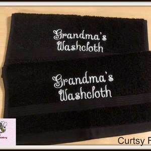 Może przedstawiać: Dwa czarne ręczniki do twarzy z białym haftem, na kt&oacute;rym widnieje napis "Grandma's Washcloth" w ozdobnym, pochylonym kroju pisma. Pod ręcznikami widoczny jest napis "Curtsy Font".