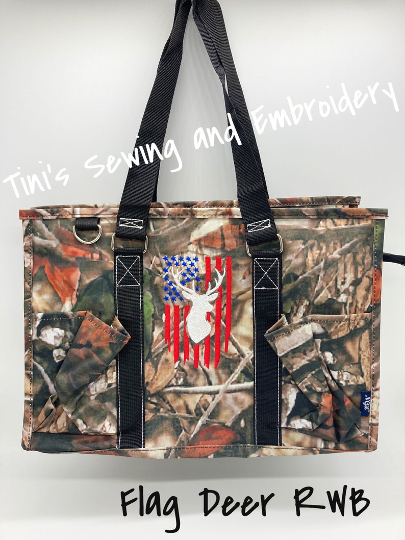 May include: A camouflage tote bag with a white deer silhouette embroidered on a red, white, and blue American flag design. The text "Flag Deer RWB" is written below the bag.
