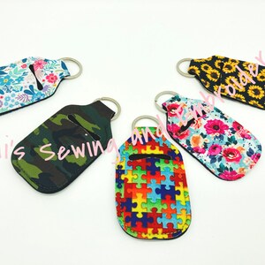 K&ouml;nnte beinhalten: F&uuml;nf bunte Handdesinfektionsmittel-Halter mit Schl&uuml;sselanh&auml;ngern. Die Halter sind aus Stoff und haben verschiedene Muster, darunter Blumen, Tarnmuster, Puzzle-Teile und Sonnenblumen.