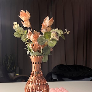 May include: A copper-coloured metal vase with a geometric design holds a bouquet of peach-coloured flowers and green leaves. The vase is sitting on a book with the text "Vogue essentials heels" on the cover. A pink candle sits on the book next to the vase.