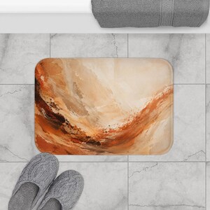 May include: A rectangular bath mat featuring an abstract painted design with swirling layers of cream, beige, burnt orange, and deep brown tones. The surface displays textured brushstroke patterns that create a fluid, layered appearance.