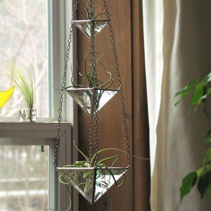 Op de afbeelding: Drie glazen driehoekige plantenhangers die aan een ketting hangen, elk met een groene luchtplant. De hangers zijn aan een raam bevestigd.