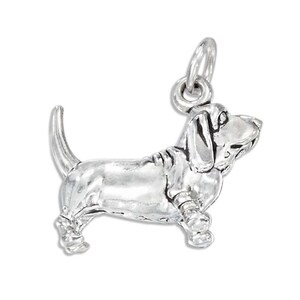 May include: A polished silver-toned metal charm shaped like a Basset Hound dog in a standing position with its head turned slightly upward. The charm features a small circular jump ring attached to the top for hanging on a chain or bracelet.