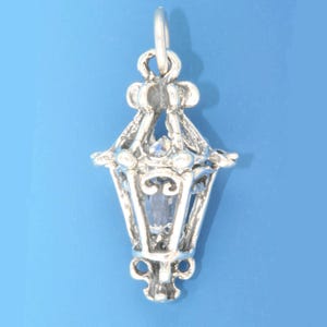 Puede incluir: Un charm de plata con forma de linterna con un cristal transparente en el centro. La linterna tiene un diseño detallado con intrincados adornos.