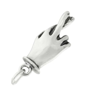 Puede incluir: Un dije de metal plateado con forma de mano con los dedos índice y medio cruzados. El dije presenta un acabado pulido con áreas hundidas oscurecidas para definir los dedos y la palma, e incluye una pequeña anilla circular en la base.