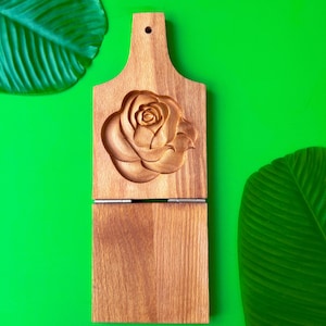 May include: A wooden cutting board with a carved rose design. The board is made of light brown wood and has a handle with a hole for hanging. The bottom section is hinged. The background is bright green.