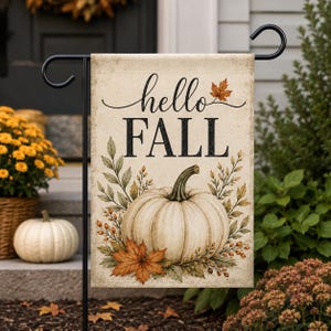 Puede incluir: Una bandera de jardín rectangular colgada en un soporte de metal negro. La bandera presenta un fondo color crema con el texto "hello FALL" en letras cursivas y de imprenta negras. Una calabaza blanca grande está centrada con hojas de otoño.