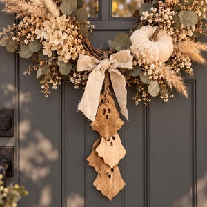 May include: A circular wreath featuring dried beige flowers, eucalyptus leaves, pampas grass, and a white pumpkin. A light beige fabric bow hangs from the center, with three wooden ghost-shaped ornaments suspended vertically from the bow's knot.