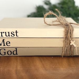 以下が含まれることがあります： クリーム、タン、トープ色の装飾的な本が積み重ねられ、紐と小さな木の十字架で結ばれています。「Trust Me -God」と黒い文字で書かれています。素朴で宗教的なテーマの家の装飾アイテム。