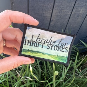 May include: A black and white patch with the text "I brake for thrift stores" and a scenic road image. The patch is held in a hand against a green grassy background.