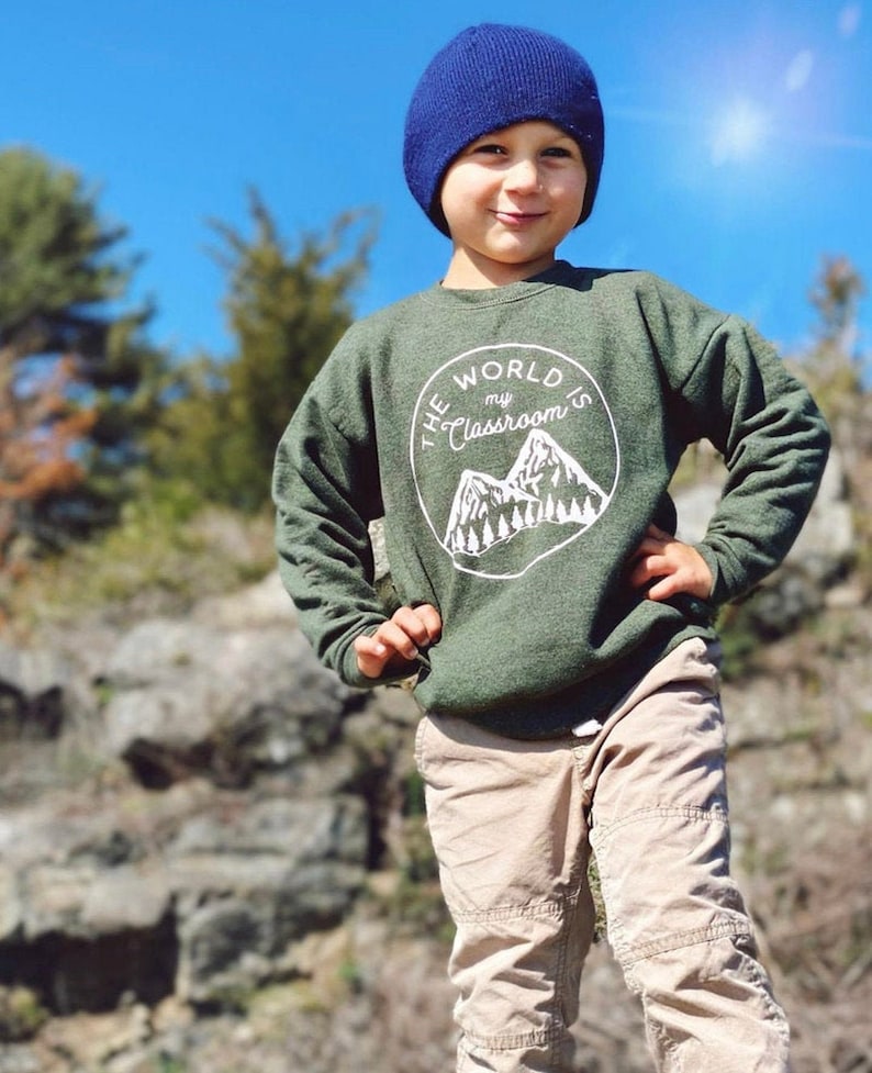 May include: A young boy wearing a green sweatshirt with the text "The World's My Classroom" in a white circle with a mountain range graphic. He is wearing a blue beanie and khaki pants. He is standing in front of a rocky background.
