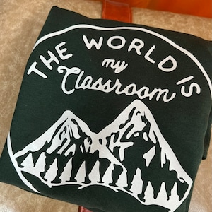 May include: A dark green sweatshirt with a white circle graphic. The graphic says "The World's My Classroom" with a mountain range and people hiking in the background.