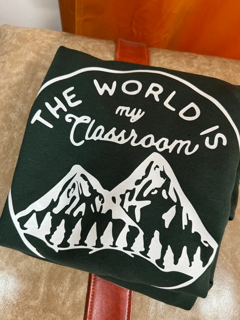May include: A dark green sweatshirt with a white circle graphic. The graphic says "The World's My Classroom" with a mountain range and people hiking in the background.