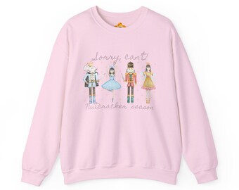 Op de afbeelding: Een lichtroze sweatshirt met een ontwerp van het Notenkrakerseizoen. De afbeelding bevat illustraties van balletpersonages, met de tekst "Sorry, Can't!" erboven en "Nutcracker season" eronder.