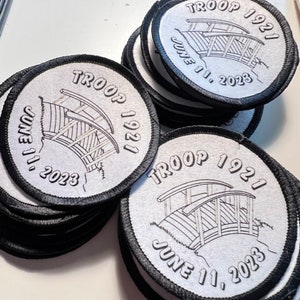 Puede incluir: Un montón de parches redondos blancos con bordes negros. Cada parche presenta el texto "TROOP 1921" y "JUNE 11, 2023" alrededor de una ilustración de un puente. Los parches son probablemente para un grupo de exploradores.