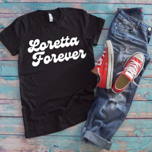 May include: Black t-shirt with white text that reads "Loretta Forever" in a retro font. The shirt is laid out on a blue wooden surface with a pair of blue jeans and red sneakers.