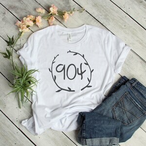May include: White t-shirt with a black graphic of a wreath with the number "904" inside. The shirt is laid flat on a wooden surface with a pair of blue denim shorts and a potted plant.
