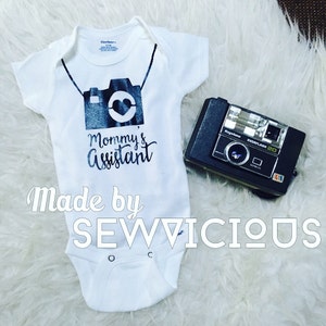 May include: White baby onesie with a black and white camera graphic and the text "Mommy's Assistant". The onesie has a white background and the text is in a black font. The onesie is made by Sewvicious.
