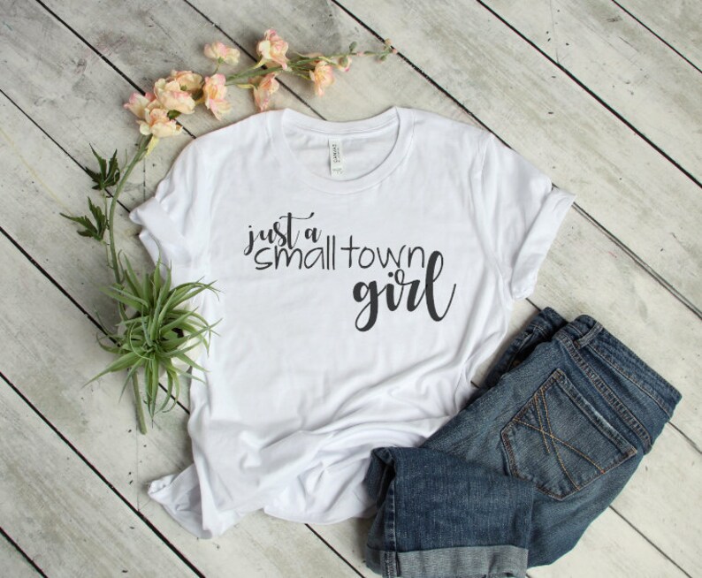 May include: White t-shirt with the text "just a small town girl" in black script font. The shirt is laid flat on a wooden surface with a pair of blue jeans.