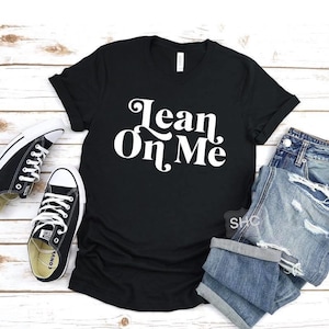 May include: Black t-shirt with white text that reads "Lean On Me". The shirt is shown with a pair of blue jeans and black sneakers.