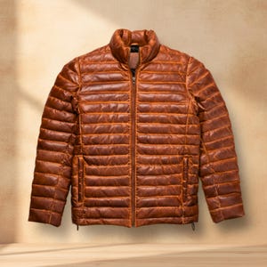 Puede incluir: Chaqueta acolchada de cuero marrón con cuello alto y cremallera completa. La chaqueta tiene costuras acolchadas horizontales y dos bolsillos laterales. La chaqueta se muestra sobre un fondo neutro.