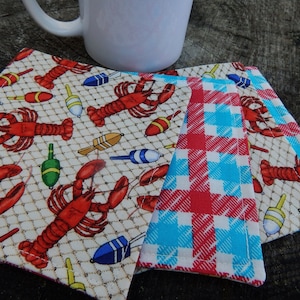 Puede incluir: Tres posavasos de tela con tem&aacute;tica n&aacute;utica. Un posavasos presenta un patr&oacute;n de cuadros rojos y azules, mientras que los otros dos posavasos presentan un fondo blanco con un patr&oacute;n de langosta y boya de pesca.