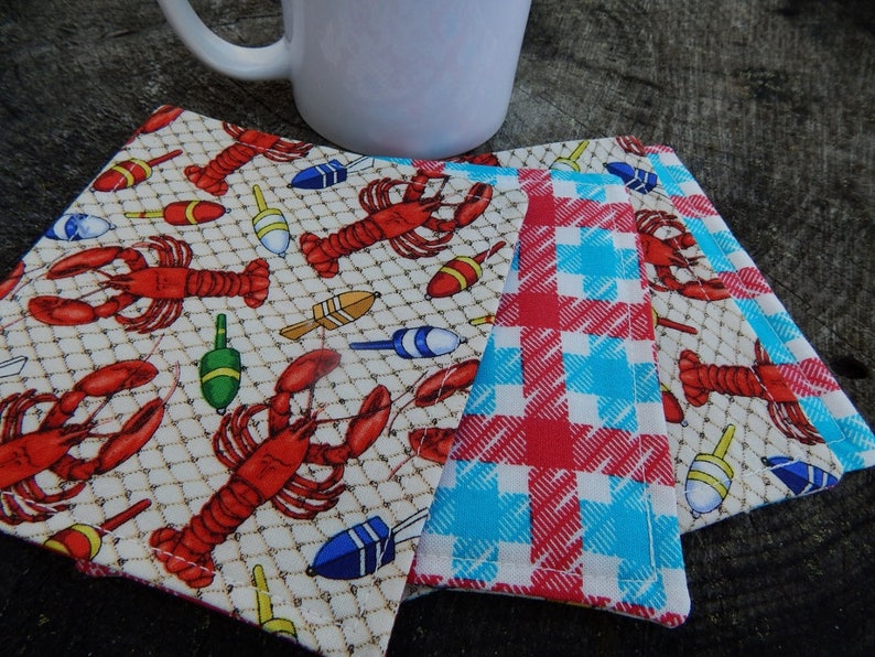 Puede incluir: Tres posavasos de tela con tem&aacute;tica n&aacute;utica. Un posavasos presenta un patr&oacute;n de cuadros rojos y azules, mientras que los otros dos posavasos presentan un fondo blanco con un patr&oacute;n de langosta y boya de pesca.