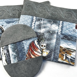 May include: A quilted nautical-themed kitchen set featuring a gray pot holder and two square hot pads. The fabric depicts a harbor scene with sailboats, ropes, and a gray wave pattern.